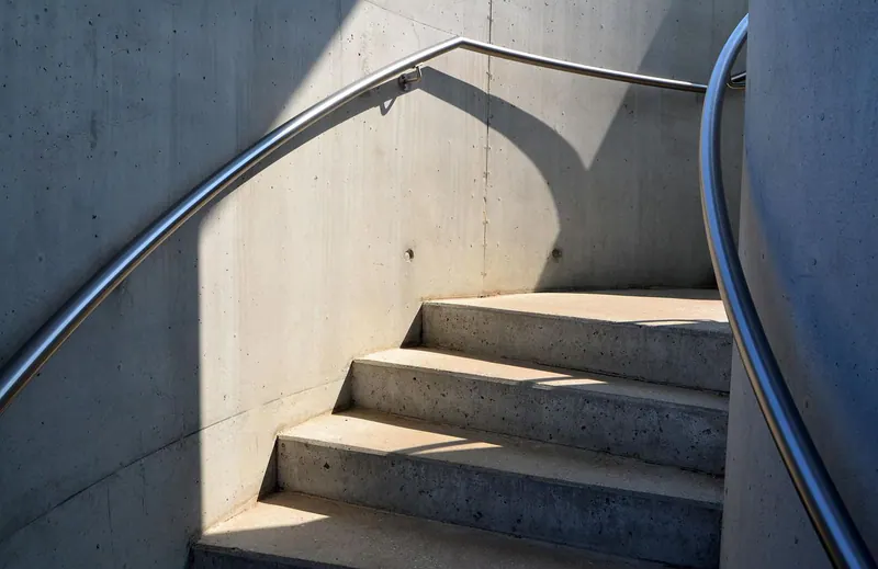Betontreppe mit geschwungenem Edelstahlhandlauf im weichen Seitenlicht, Schattenspiel auf der Sichtbetonwand