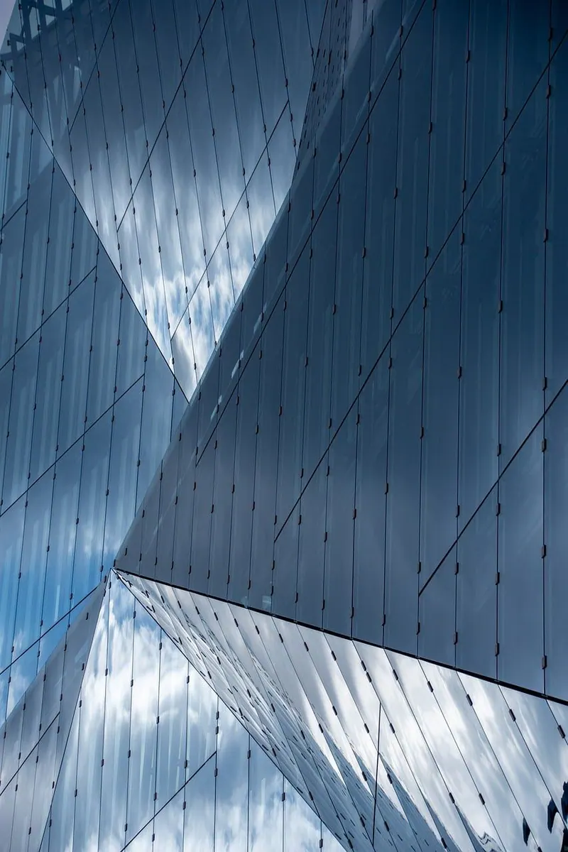 Detailaufnahme einer facettierten Glas- und Metallfassade mit Wolkenspiegelung und präzisen Fugen